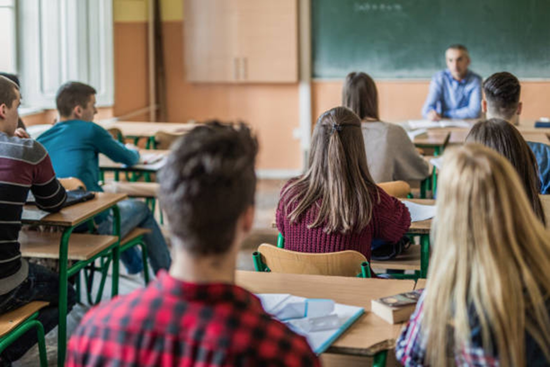 An over the shoulder shot of students sat at desks being taught by a teacher at the front desk