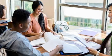 Smiling students sat at a table together looking at oneanother and their work